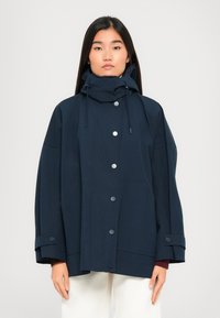 Woman wearing a navy oversized hooded jacket with snap buttons and adjustable cuff straps, paired with white pants, standing against a plain background.