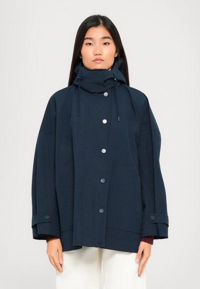 Egy nő tengerészkék, oversized kapucnis kabátban áll, amely patentgombokkal és állítható mandzsettákkal rendelkezik, fehér nadrággal párosítva, egy sima háttér előtt.