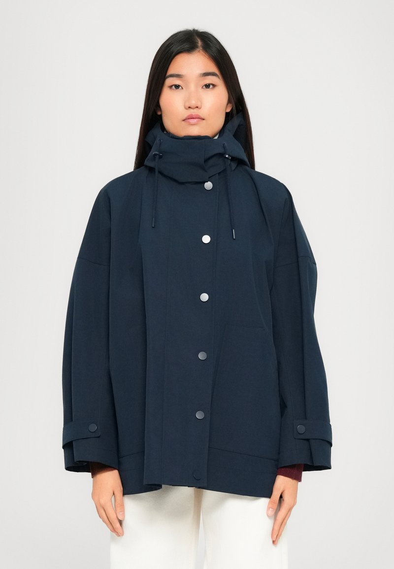 Woman wearing a navy oversized hooded jacket with snap buttons and adjustable cuff straps, paired with white pants, standing against a plain background.