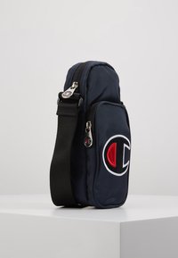 Granatowa torba typu crossbody z zamkiem błyskawicznym, regulowanym czarnym paskiem oraz haftowanym logo z czerwonymi i białymi akcentami z przodu.