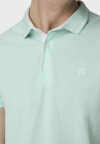 Hellgrünes Poloshirt mit strukturiertem Stoff, zwei Knöpfen und einem weißen Kragen. Mit einem kleinen weißen Logo auf der Brust.