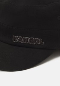 Cappello di cotone nero con forma arrotondata e visiera cucita. Presenta il testo ricamato "KAN GOL" in grigio. Tessuto testurizzato con una finitura liscia.