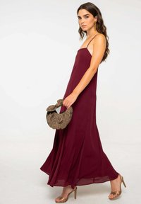 Robe maxi bordeaux avec des bretelles spaghetti, en tissu fluide et avec un léger éclat. Associée à un sac à main en cuir marron texturé et des talons compensés dorés.