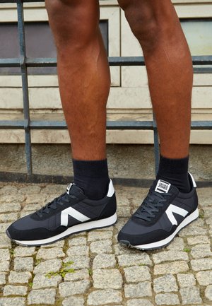 Scarpe da ginnastica nere con accenti bianchi, tessuto testurizzato e design elegante. Presentano lacci e un logo sulla linguetta. Indossate con calzini neri alla caviglia.