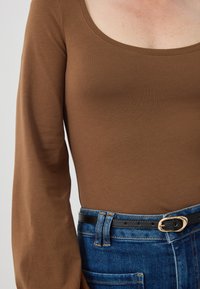 Braunes Langarmoberteil aus weichem Material mit weitem Halsauschnitt, kombiniert mit hoch geschnittenen blauen Jeans und einem schwarzen Gürtel mit einer goldenen Schnalle.