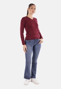 Top lungo a maniche lunghe in V-bordeaux, silhouette aderente, abbinato a jeans blu a zampa d'elefante e sneakers bianche. Design semplice, texture liscia.