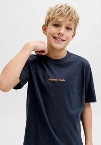 Ξανθός αγόρι που χαμογελάει, φοράει σκούρο μπλε μπλουζάκι με το κείμενο "Originals Studio", σηκώνοντας το δεξί του χέρι κοντά στον ώμο του, με φόντο απλό.