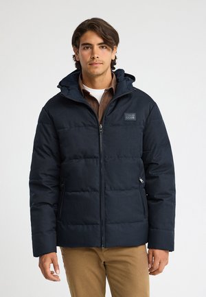Joven con una chaqueta acolchada azul marino con capucha, con cremallera, sobre una camisa marrón y pantalones beige, de pie frente a un fondo liso.