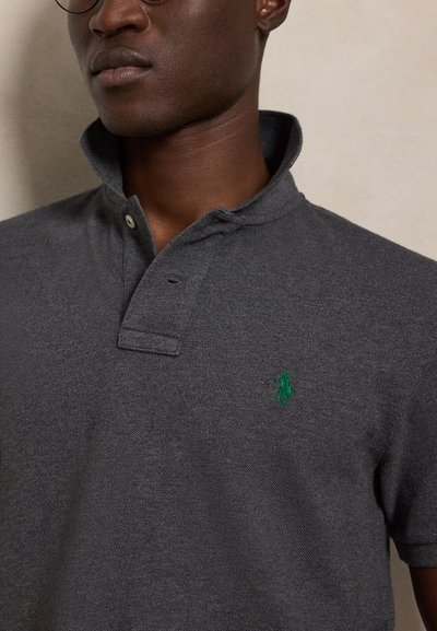 Homme portant un polo gris foncé avec un col relevé et un logo vert brodé sur la poitrine.