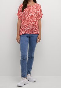 Blusa de patrón floral rojo y blanco con mangas cortas, combinada con jeans ajustados azules y zapatillas blancas. Ligera, de corte relajado.