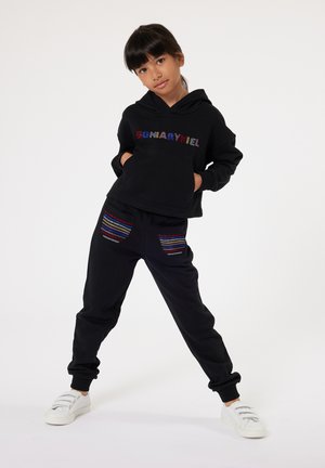 Haut à capuche noir avec le texte "SONARYHEL" en perles colorées, associé à un pantalon noir avec des accents de poches rayés. Des baskets blanches complètent la tenue.