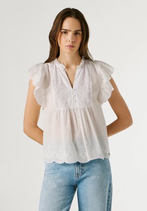 Mujer joven con blusa blanca de encaje ojete con mangas cortas fruncidas y jeans azul claro, de pie contra un fondo liso.