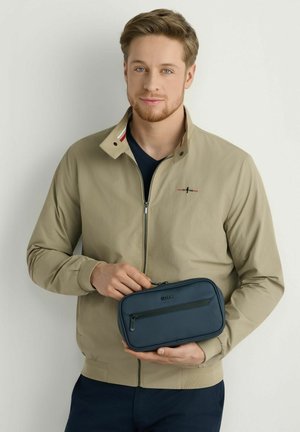 Młody mężczyzna w beżowej kurtce trzyma granatową saszetkę na suwak z przednią kieszenią na jednolitym tle.