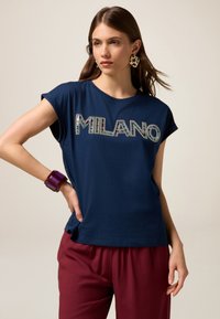 Tricou din bumbac de culoare albastru marin, cu mâ sleeves scurte, având inscripția „MILANO” din strasuri multicolore. Asortat cu pantaloni burgundy și o brățară voluminoasă.