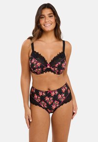Femme souriante portant un soutien-gorge en dentelle noire florale et un slip taille haute assorti, se tenant devant un fond clair uni.
