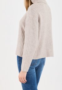 Pull côtelé beige clair avec un col montant et des manches longues, doté d'un ourlet court. Le tissu semble doux et texturé. Porté avec un jean bleu.