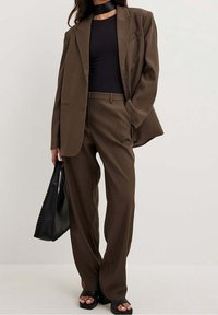 Brązowy garnitur szyty na miarę z luźną marynarką i szerokimi spodniami, połączony z czarnym topem i dużą czarną torebką. Całość dopełniają czarne sandały na platformie.