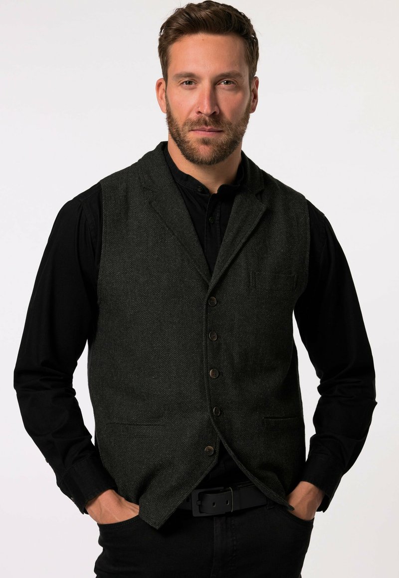 Donkergrijze herringbone vest met vijf knopen, notch revers en één borstzak, gedragen over een zwart langslijf shirt.