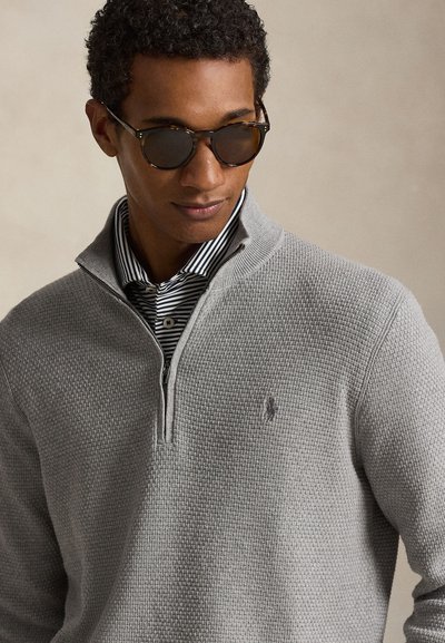 Uomo che indossa occhiali da sole tartarugati, maglione grigio testurizzato con zip a quarto e camicia a righe bianche e nere con collo, guarda verso il basso.