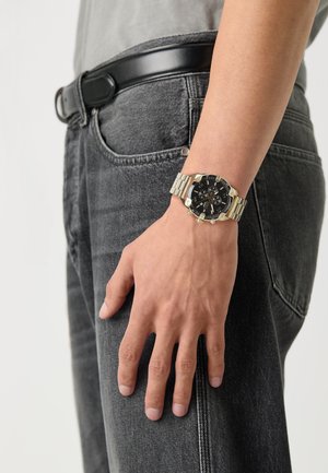 Sort urskive armbåndsur med sølv rustfrit stålbånd, der har et chronograf-design og guld accenter, båret på en hånd over grå jeans.