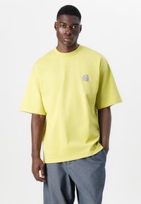 T-shirt en coton jaune à manches courtes, col rond, avec un détail logo violet sur la poitrine gauche. Coupe ample, texture unie.