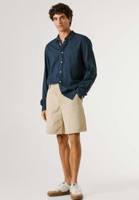 Junger Mann trägt ein marineblaues Hemd mit Knöpfen, beige knielange Shorts und weiße Sneakers, steht vor einem schlichten Hintergrund.