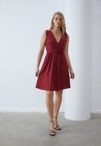 Punainen polvipituinen mekko, jossa on kietaisusuunnittelu, hihattomat malli ja syvä V-kaula-aukko. Silkkinen kangas, jossa on kerätty vyötärödetail. Mustat nauhakenkä.