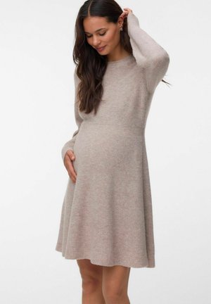 Gravid kvinde med langt mørkt hår iført en beige strikket kjole, der blidt holder sin mave og rører ved sit hår, stående mod en hvid baggrund.