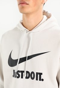 Ljus beige hoodie i bomullsmix, med en svart Nike-logotyp och texten "JUST DO IT." Justerbar snörning i huvan.