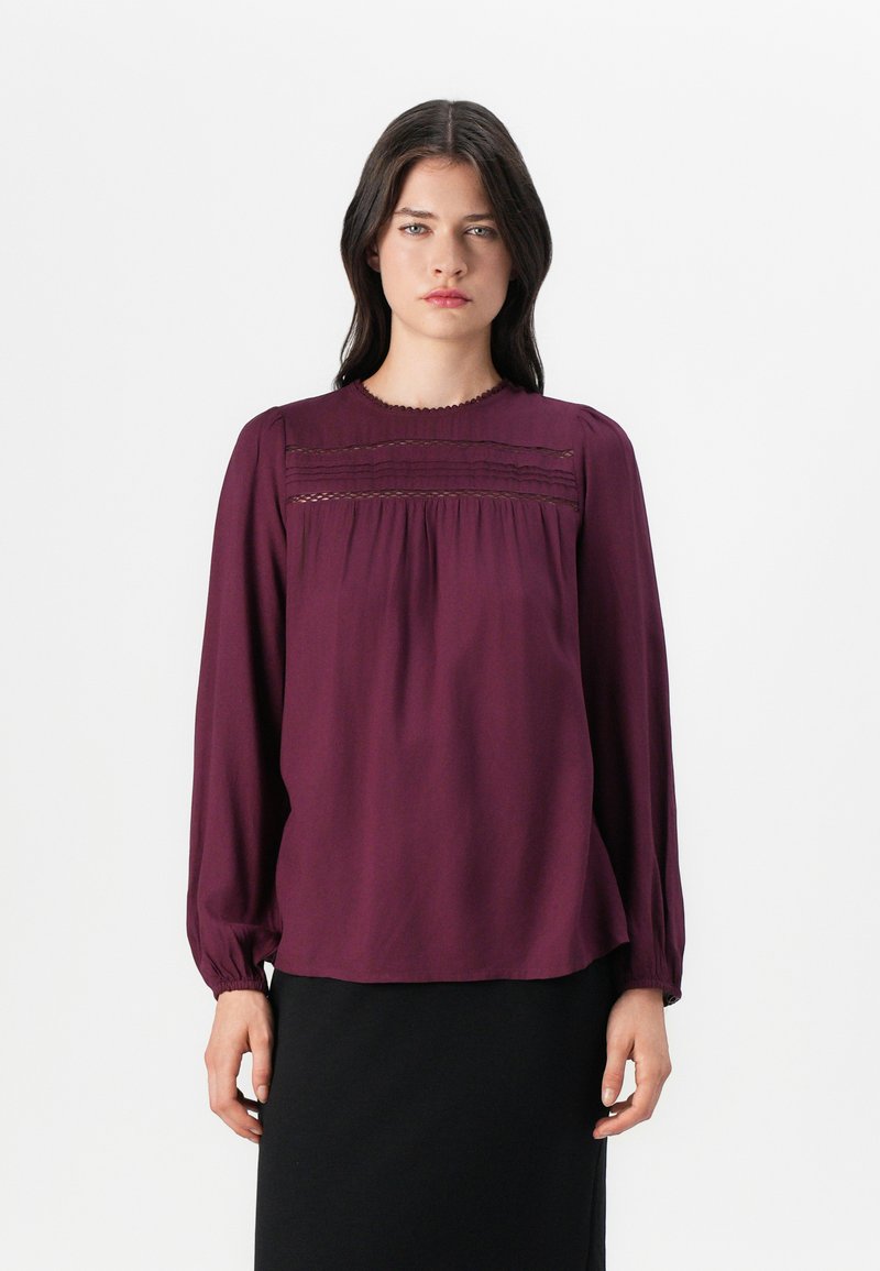 Burgundy blus i lätt tyg, med långa ärmar, rynkdetaljer vid bodyn och en klassisk rund halsringning.