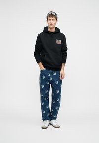 Musta huppari graafisella yksityiskohdalla, yhdistettynä sinisiin farkkuihin, joissa on yin-yang-kuvio. Valkoiset ja harmaat lenkkarit täydentävät asukokonaisuuden.