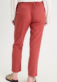 Personne portant un pantalon couleur rouille avec une taille élastique, une chemise blanche à manches longues et des sandales beiges à enfiler avec des détails décoratifs.