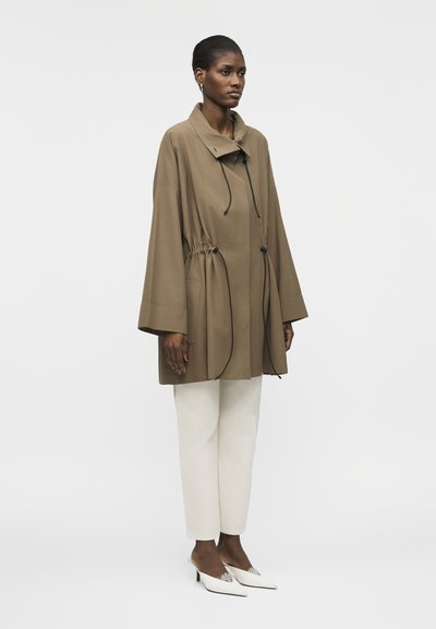 Žena v nadrozmernej hnedej bunde so šnúrkou v páse, bielych rovnomerných nohaviciach a bielych špičatých lodičkách, stojaca pred jednoduchým bielym pozadím.