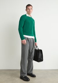 Pulover verde tricotat peste o cămașă albă cu guler, pantaloni gri și pantofi negri. Ține o geantă tote texturată neagră. Fundal neutru.