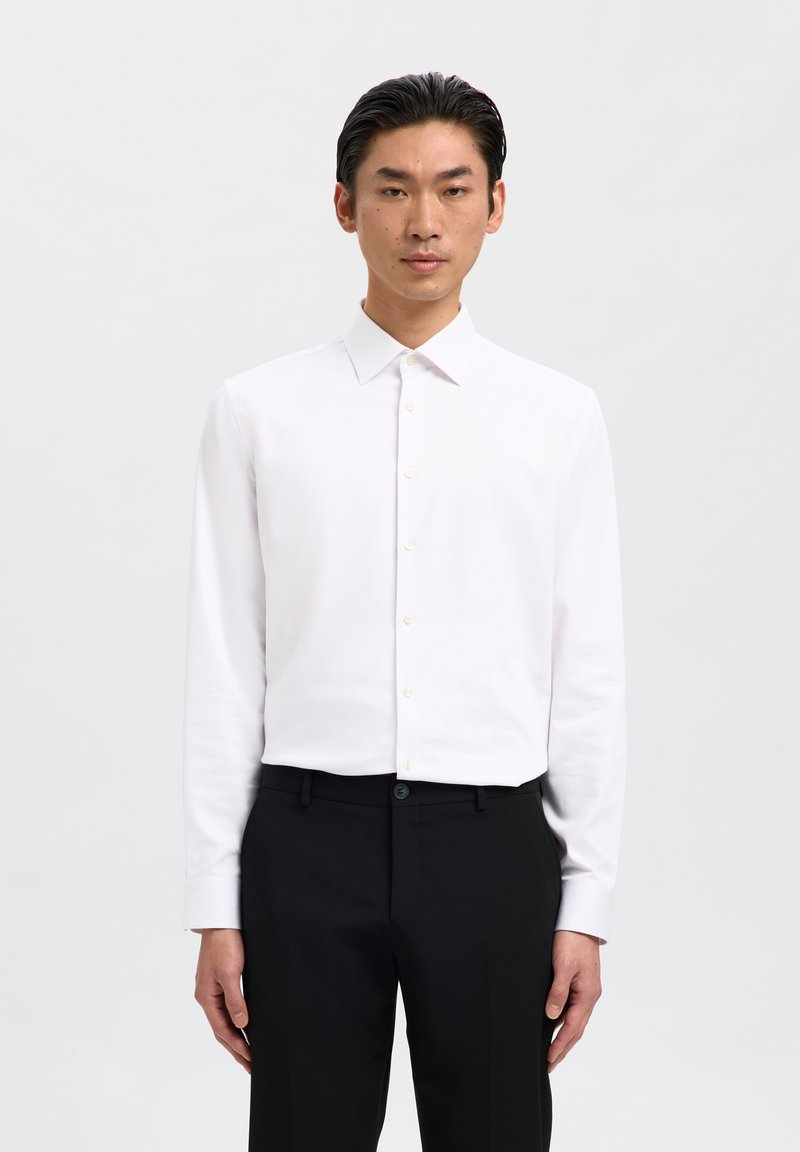 Hombre con una camisa blanca entallada abotonada y pantalones negros, de pie frente a un fondo blanco liso, mirando hacia adelante.