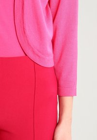 Růžový pletený svetr s kulatým límcem a dlouhými rukávy, v kombinaci s červenými kalhotami na míru s texturovaným povrchem.