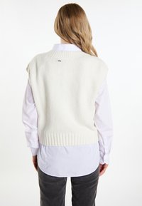 Gilet bianco a maglia con orlo a costine, indossato sopra una camicia bianca con bottoni. La texture sembra morbida, senza motivi o accenti visibili.