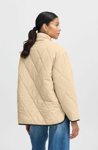 Veste matelassée beige avec un col montant et des finitions noires, présentant une coupe décontractée et un motif de piqûre en losange. Portée avec un jean bleu à jambes larges.