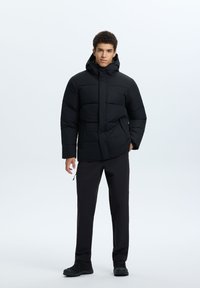 Zwarte pufferjas met een capuchon, voorzien van een doorgestikte ontwerp en een ritssluiting aan de voorkant. Gekoombineerd met zwarte broek en laarzen, staand tegen een neutrale achtergrond.