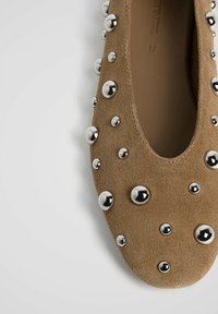 Chaussure plate en daim marron décorée de clous argentés brillants de différentes tailles sur fond blanc.