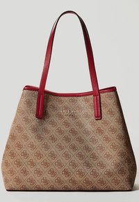 Beige kassi, jossa on teksturoitu pinta, punaiset synteettisestä nahasta valmistetut kahvat, hienovarainen logotyyppi ja geometrinen kuvio koko kassin pinnalla.