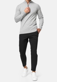 Maglione grigio con zip e orlo a coste, indossato con pantaloni neri a gamba stretta e sneakers bianche. Tessuto a maglia morbido, design aderente.