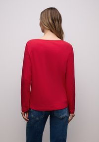 Vrouw met lang haar, gekleed in een rood shirt met lange mouwen en een blauwe spijkerbroek, achterwaarts gericht tegen een effen lichte achtergrond.