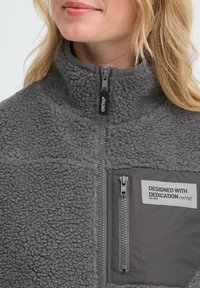 Femme portant une veste en polaire grise avec une fermeture éclair frontale et une poche sur la poitrine arborant une étiquette qui dit "CONÇU AVEC DÉVOUEMENT oxmo®".