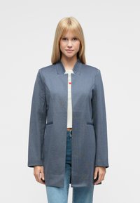 Dlhý sivý kabát s golierom z tkaného materiálu. Má bočné vrecká a rovný strih. Nosený cez bielu košeľu s červeným srdcovým motívom.