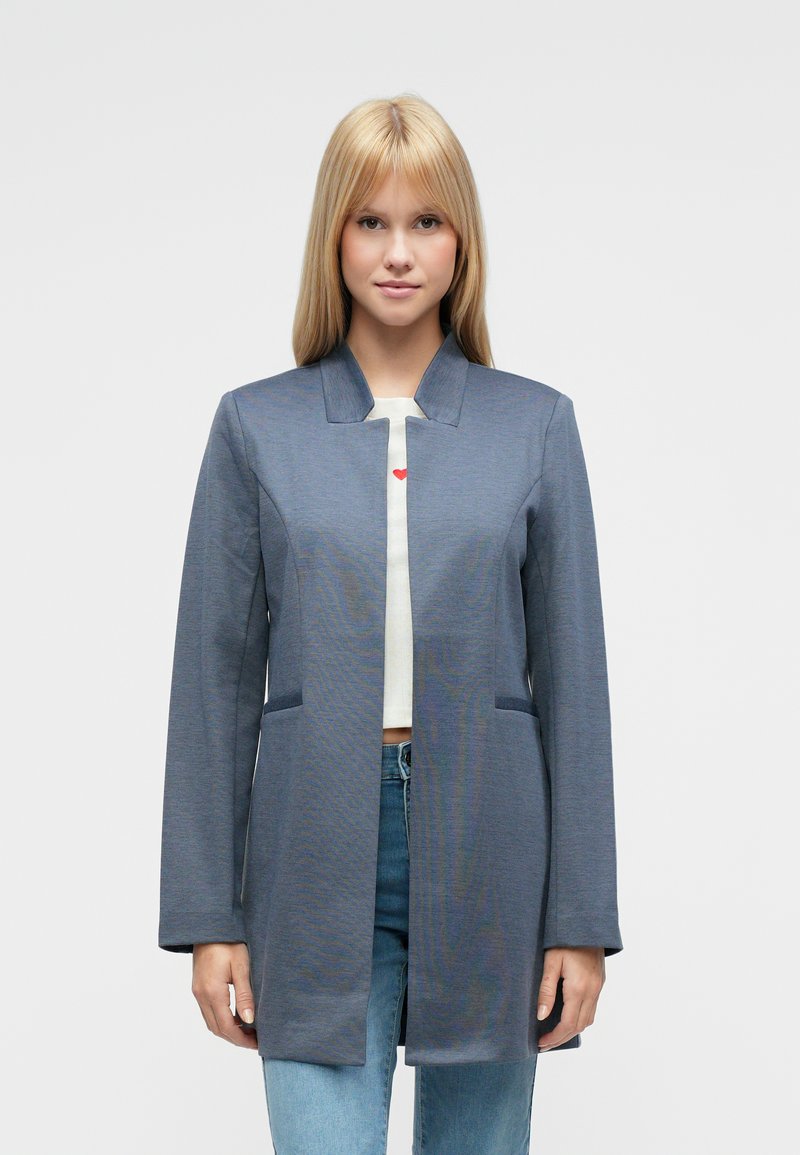 Dlhý sivý kabát s golierom z tkaného materiálu. Má bočné vrecká a rovný strih. Nosený cez bielu košeľu s červeným srdcovým motívom.