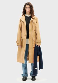 Beige Trenchcoat mit großen Fronttaschen, Knopfverschluss und Kapuze. Über einem schwarzen Strickpullover, einem hellen Hemd und blauen Jeans getragen.