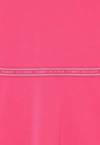 Różowa koszulka z gładką fakturą i kontrastującym szarym paskiem z napisem "TOMMY HILFIGER" powtórzonym dwukrotnie na środku.