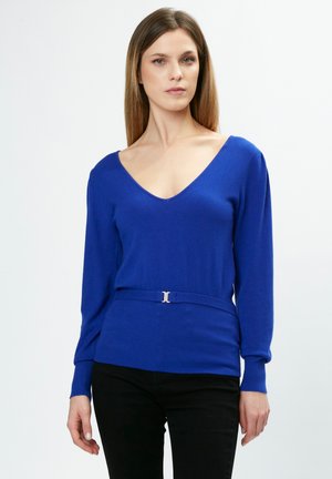 Pull bleu à manches longues avec un décolleté en V profond, taille ajustée et ceinture argentée. Fabriqué dans un tissu lisse et texturé. Pantalon noir porté en dessous.