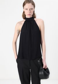 Zwart mouwloos topje met halternek, verzameld materiaal bij de halslijn, gladde textuur en een losse pasvorm, gecombineerd met zwarte broek en handtas.